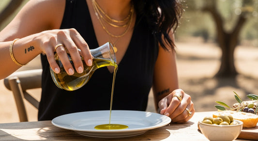 Person gießt extra natives Olivenöl aus einer klaren Glas-Karaffe auf einen Teller auf einem natürlichen Holztisch, im Hintergrund ein sonniger Olivengarten